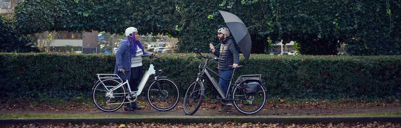 A woman and man on RadCity 5 Plus electric bikes chat in the rain while standing alongside their bikes. The man holds an umbrella.