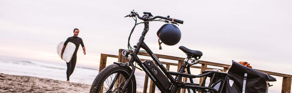 Man walks out of ocean holding longboard toward cargo ebike on beach.