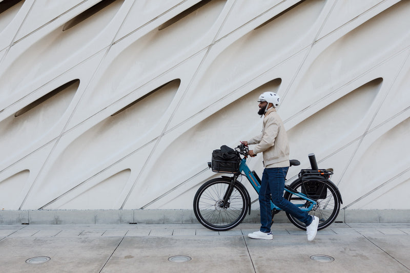 Man walking a blue Radster Road electric commuter bike in front of a building with a geometric fascade. 