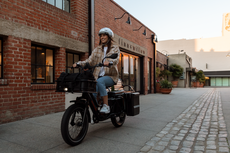 Image of a woman riding the RadWagon 5 on a sidewalk. She is wearing a helmet & the bike is equipped with a large front-mounted basket & rear-mounted hardshell panniers.