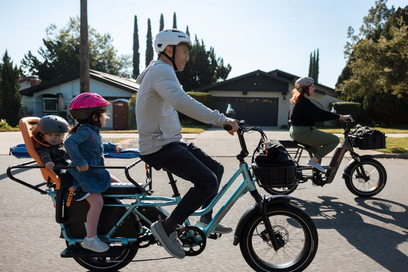 Man riding a blue RadWagon 5 electric cargo bike with two children as passengers. The children are seated with passenger accessories including the RadWagon 5 Deckpad, an orange thule yepp child seat and a caboose.