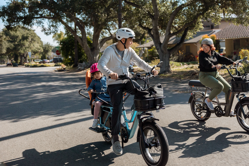An image of a man riding the RadWagon 5 in Metallic blue. His daughter is on the back of the bike in a Thule Yepp Child Seat. They are riding down a residential street. A woman is riding beside them on a RadRunner 3 Plus with a large front-mounted basket. All people are wearing helmets.