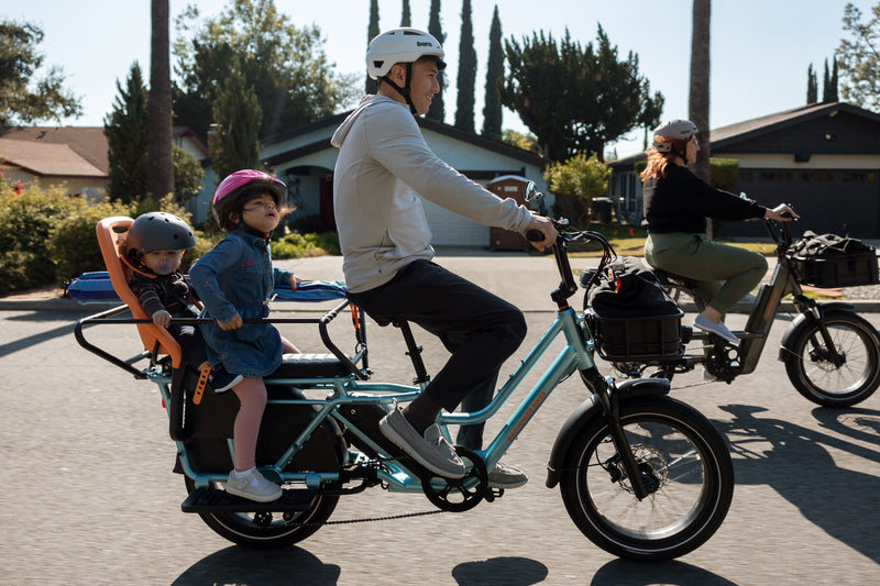 Man riding a blue RadWagon 5 electric cargo bike with two children as passengers. The children are seated with passenger accessories including the RadWagon 5 Deckpad, an orange thule yepp child seat and a caboose.