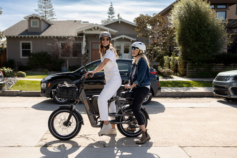 Two women riding on a RadRunner 3 Plus electric utility bike, one driving and the other sitting on the passenger seat.
